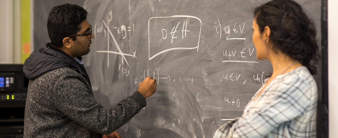 Two people working out math problem at a blackboard