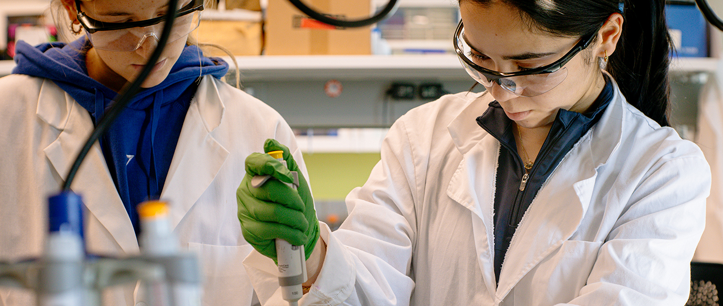 Two students work in a research lab with goggles and lab coats